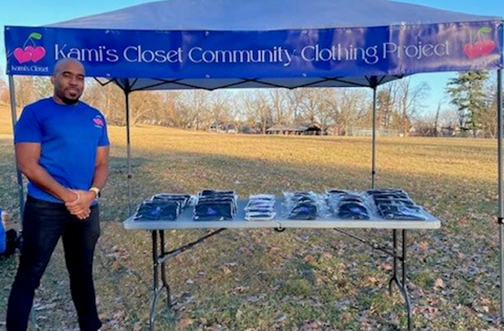 A man stands next to a table lined with winter hats, under a blue tent with a banner scribed with Kami's Closet Community Clothing Project